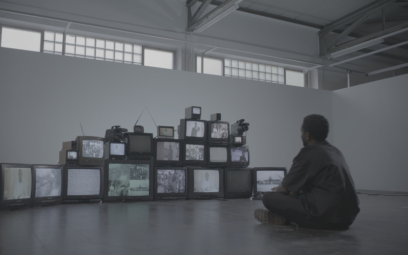 Zu sehen ist eine am Boden sitzende Person, die eine Installation aus alten Fernsehern betrachtet.
