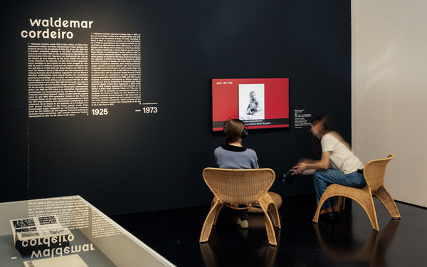 Two people are sitting in wicker chairs in front of a screen.
